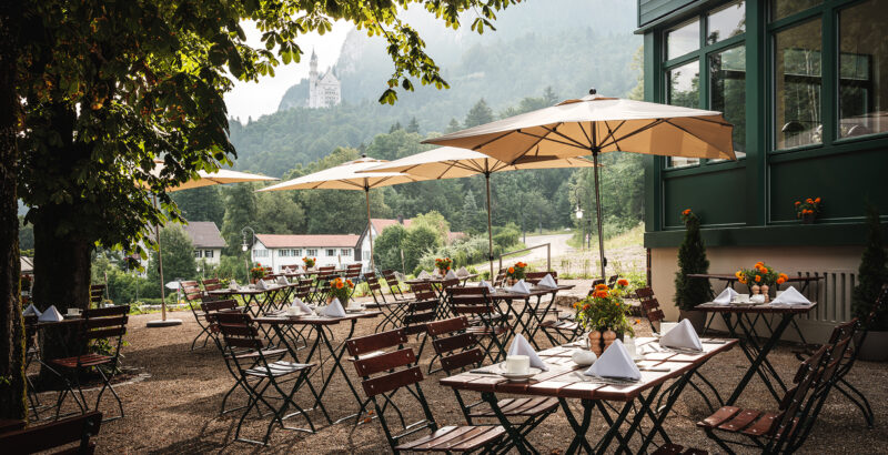 Terrasse mit mehreren Stühlen und Tischen unter Bäumen; dahinter Blick auf See und Landschaft im warmen Licht.