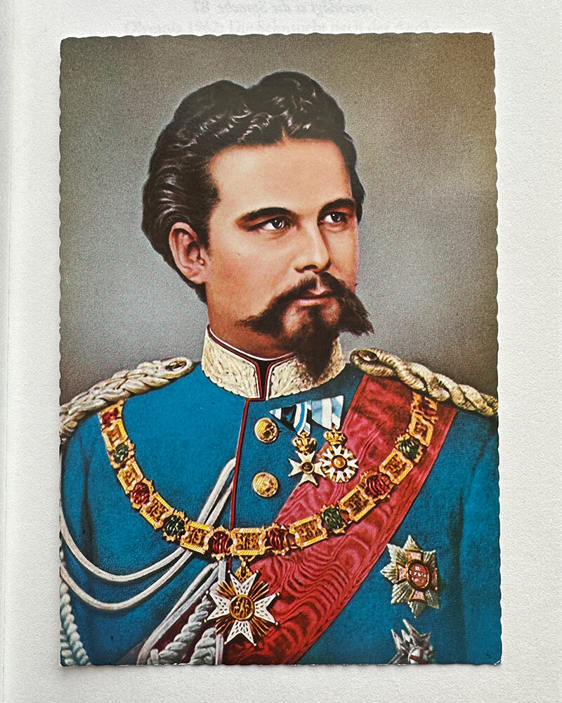 Gemälde Ludwigs II. mit dunklem Haar und Schnurrbart in blauer Uniform mit roten und goldenen Verzierungen.
