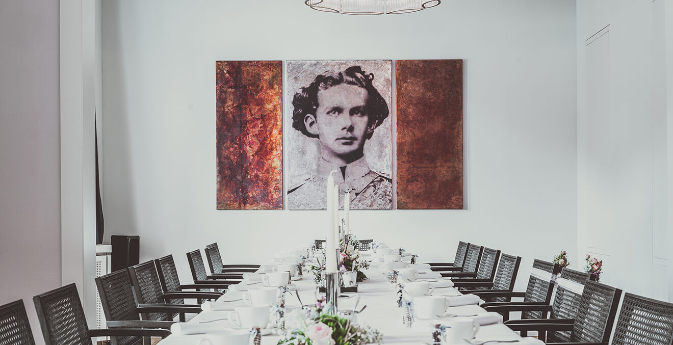 Eingedeckte Essenstafel mit weißer Tischdecke und weißem Geschirr. An der Wand ein großes Porträt von König Ludwig II.