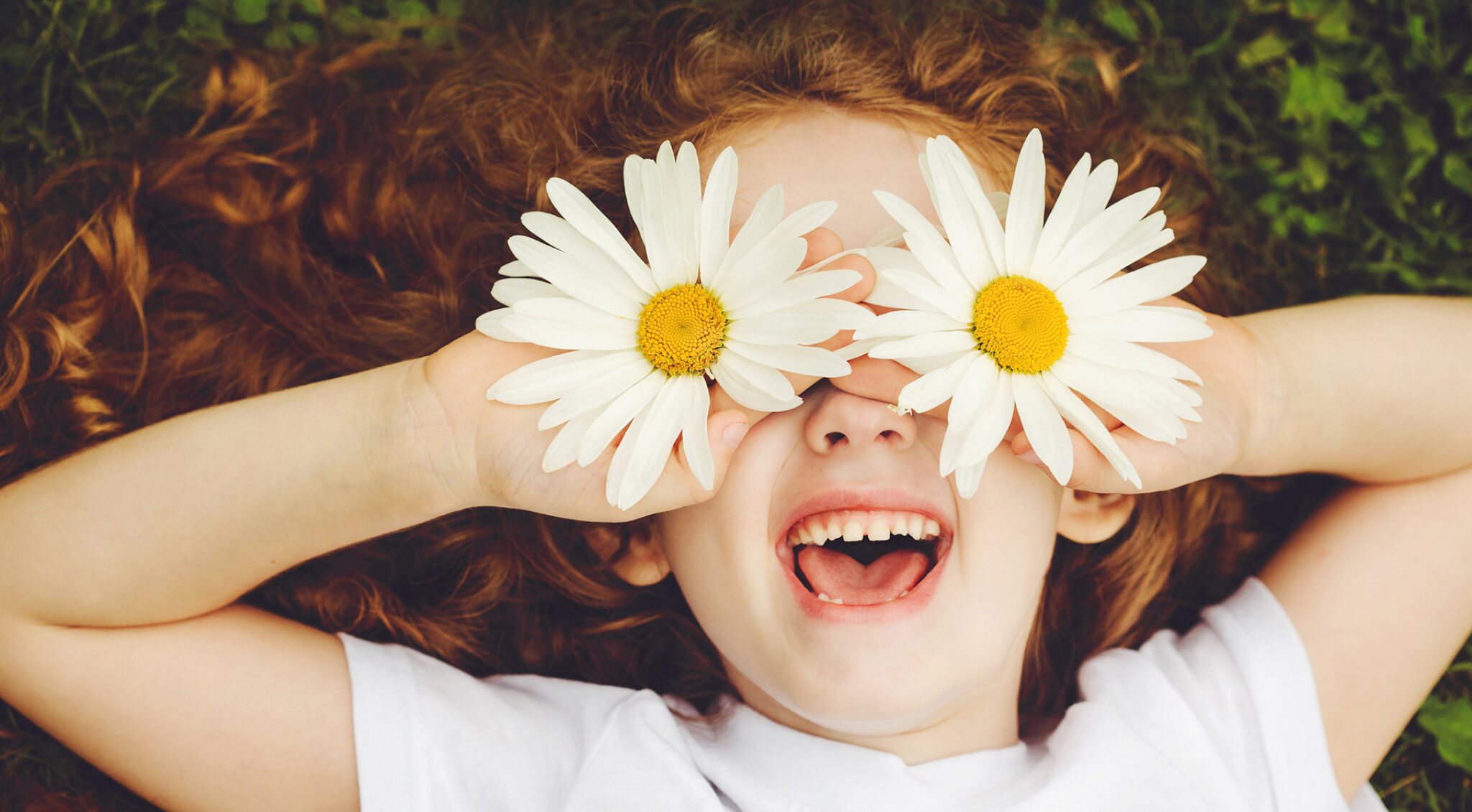 Kind mit langen rötlichen Haaren hält zwei weiße Gänseblumen vor die Augen, sodass die Blüten die Augen verdecken; das Kind lächelt und zeigt die Zähne.