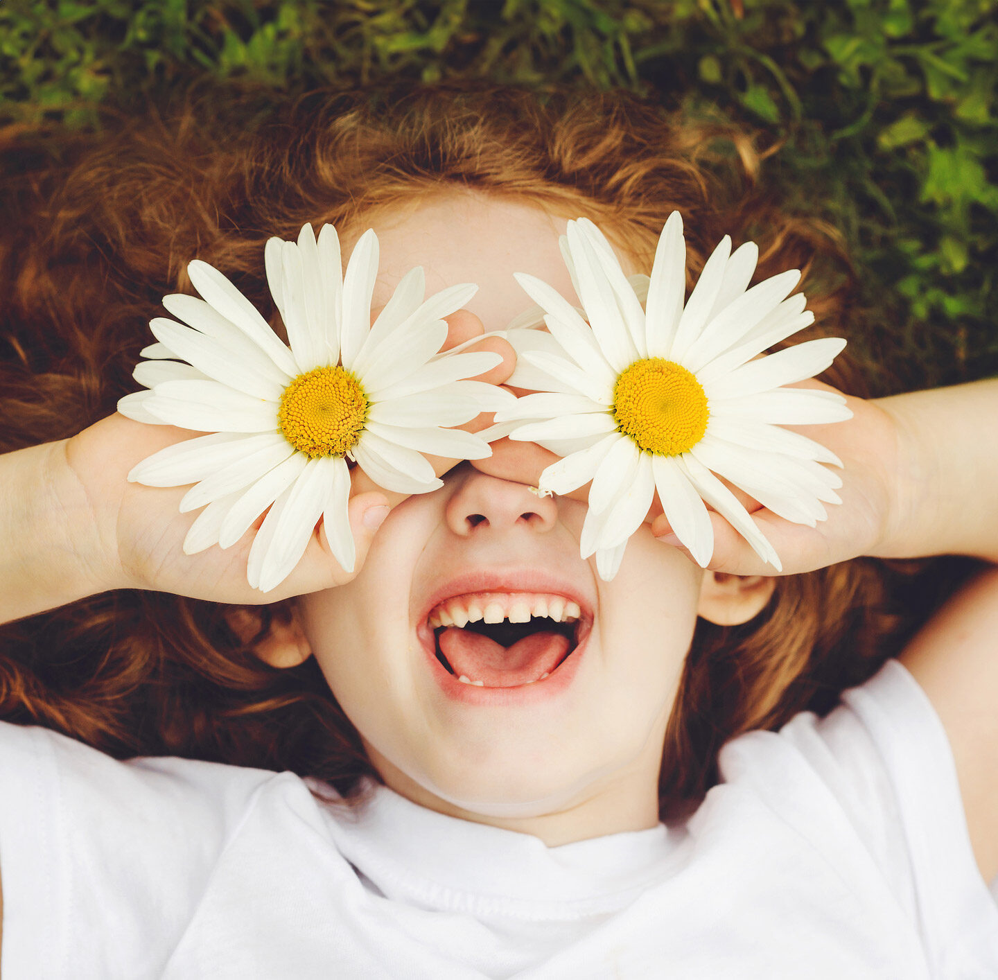 Kind mit langen rötlichen Haaren hält zwei weiße Gänseblumen vor die Augen, sodass die Blüten die Augen verdecken; das Kind lächelt und zeigt die Zähne.