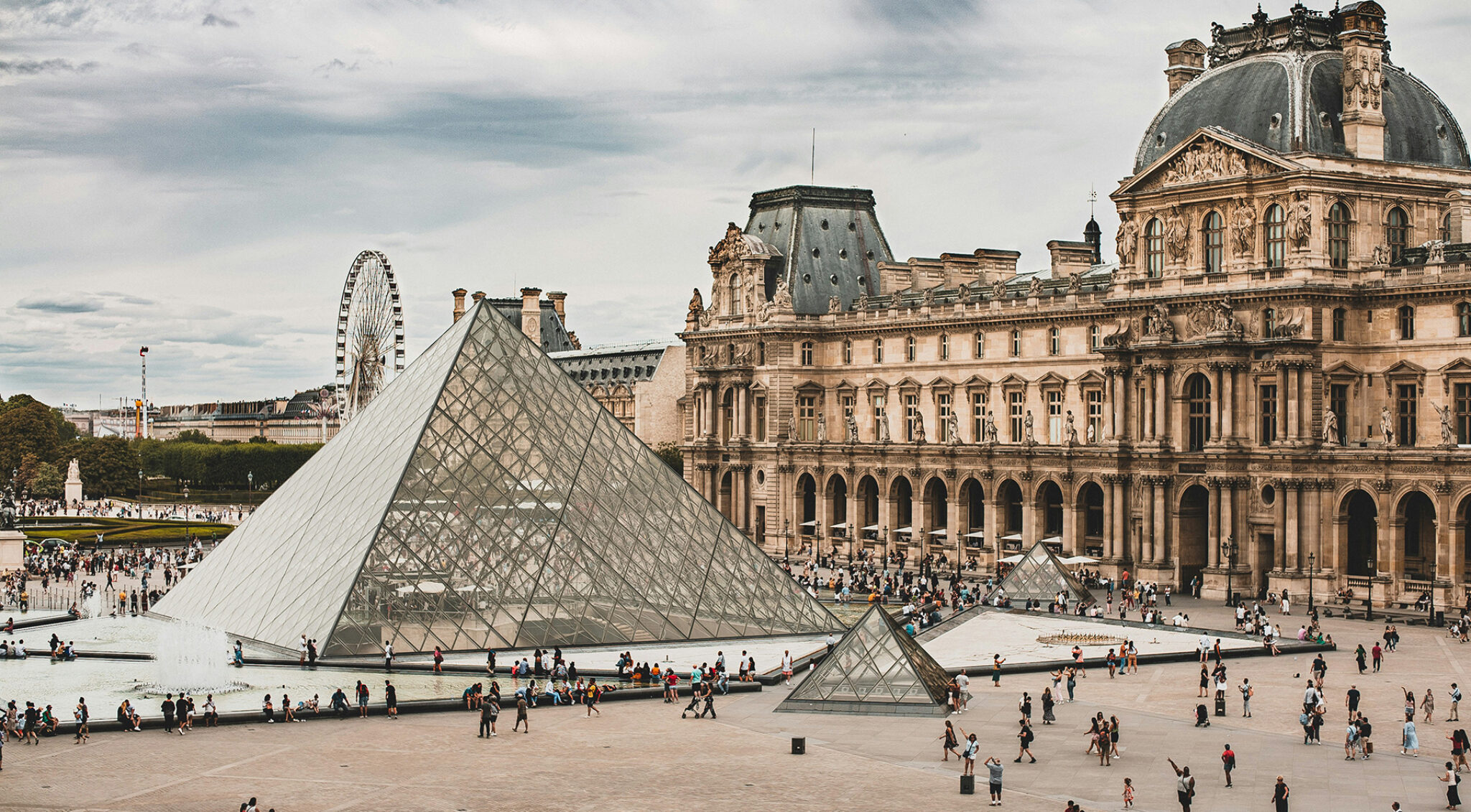 Panoramaansicht von Paris mit dem Louvre-Gebäude rechts, der Glaspyramide und einem Riesenrad links im Hintergrund unter hellem Himmel.
