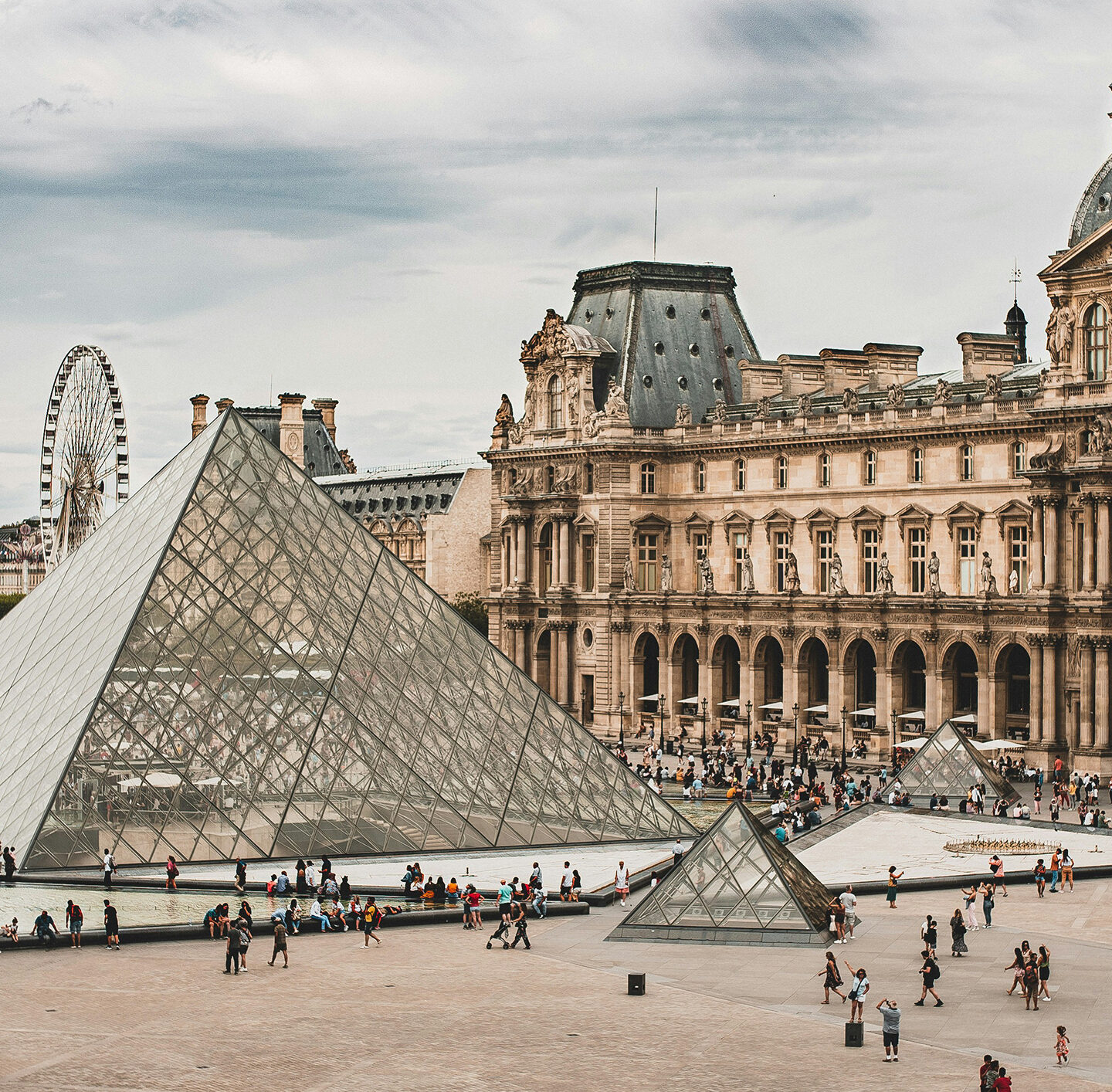 Panoramaansicht von Paris mit dem Louvre-Gebäude rechts, der Glaspyramide und einem Riesenrad links im Hintergrund unter hellem Himmel.