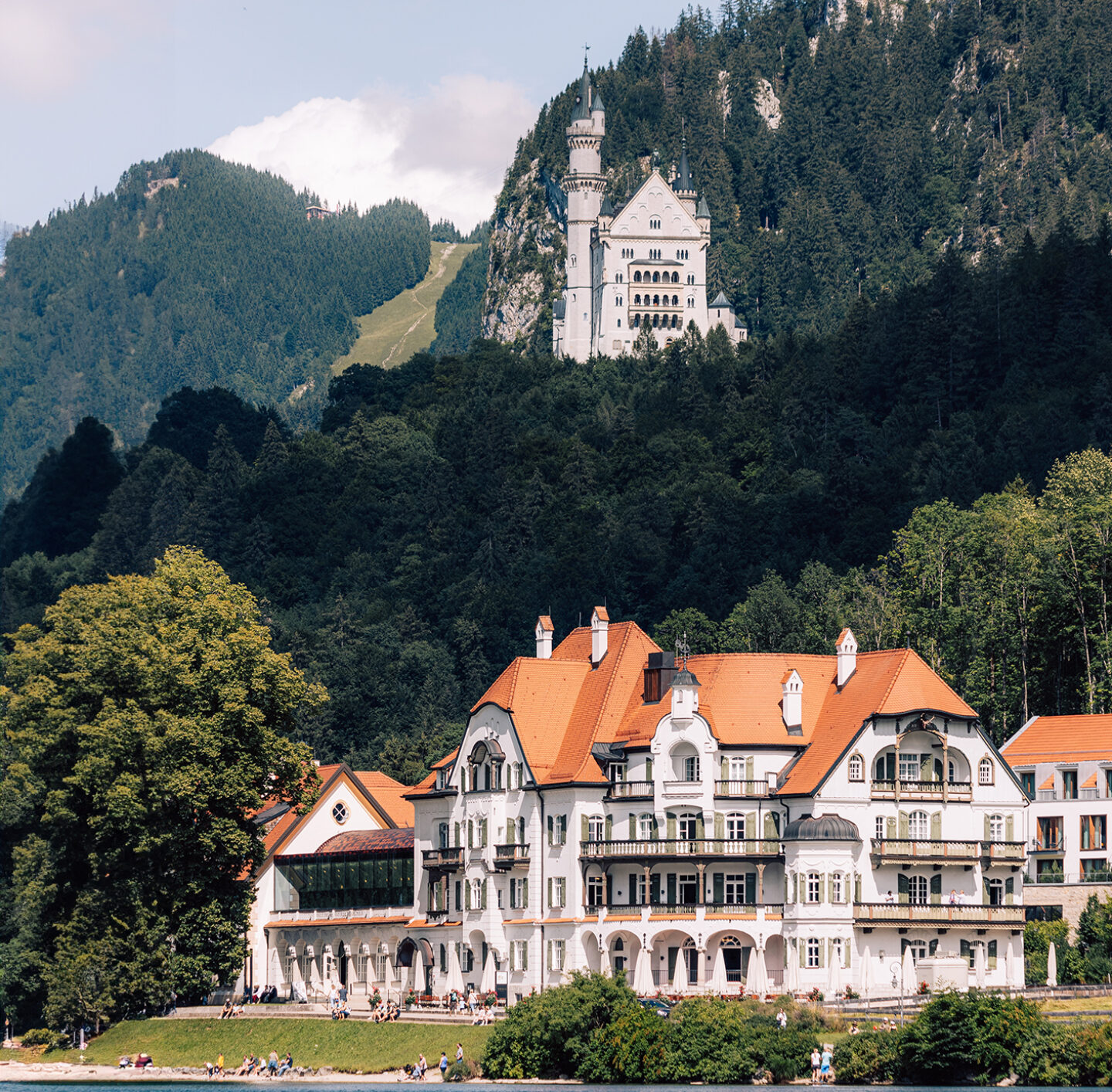 Blick über einen See auf eine weite Landschaft mit Wiesen, einzelnen Gebäuden, Schloss Neuschwanstein und bewaldeten Hügeln im Hintergrund; im Vordergrund rahmen Äste mit grünen Blättern die Szene ein.