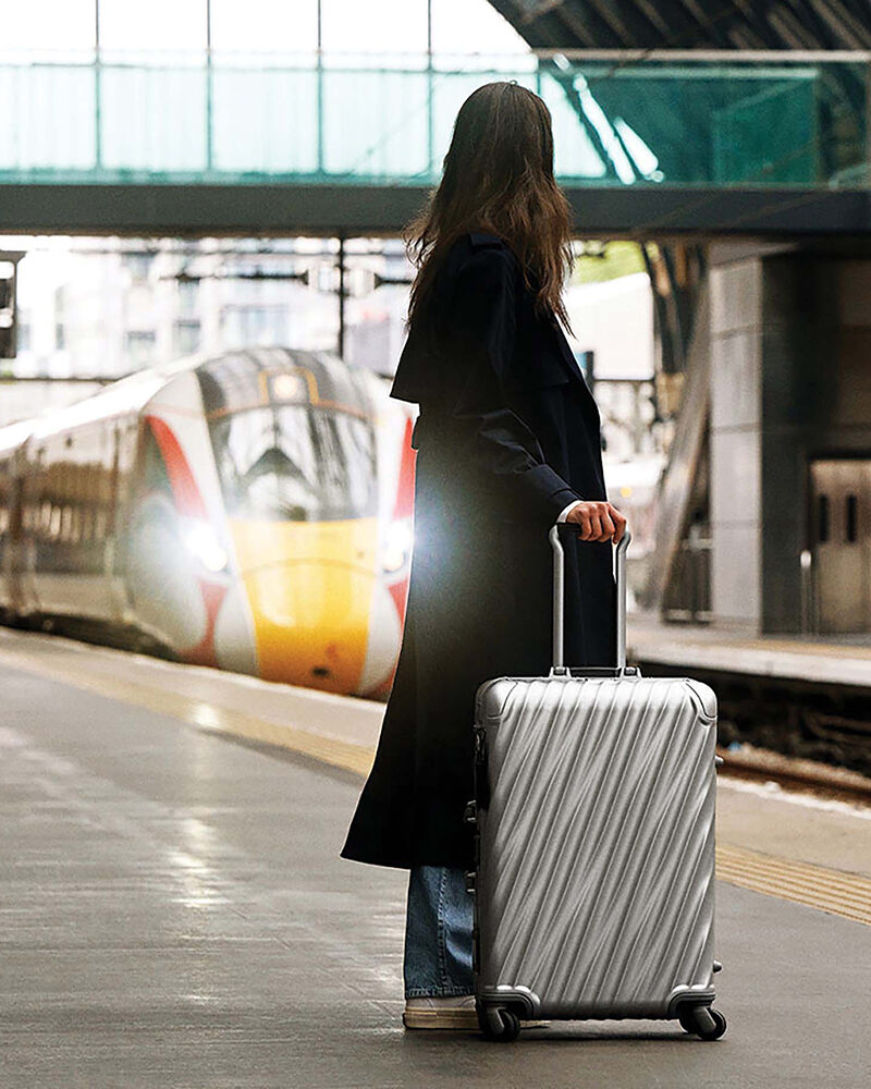 Eine Frau mit dunklem Mantel zieht einen silberfarbenen Rollkoffer auf einem Bahnhof; im Hintergrund fährt ein Zug ein.