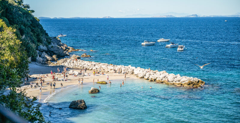 Bucht mit türkisfarbenem Wasser und hellem Sandstrand; im Vordergrund rahmen grüne Pflanzen die Küste ein.
