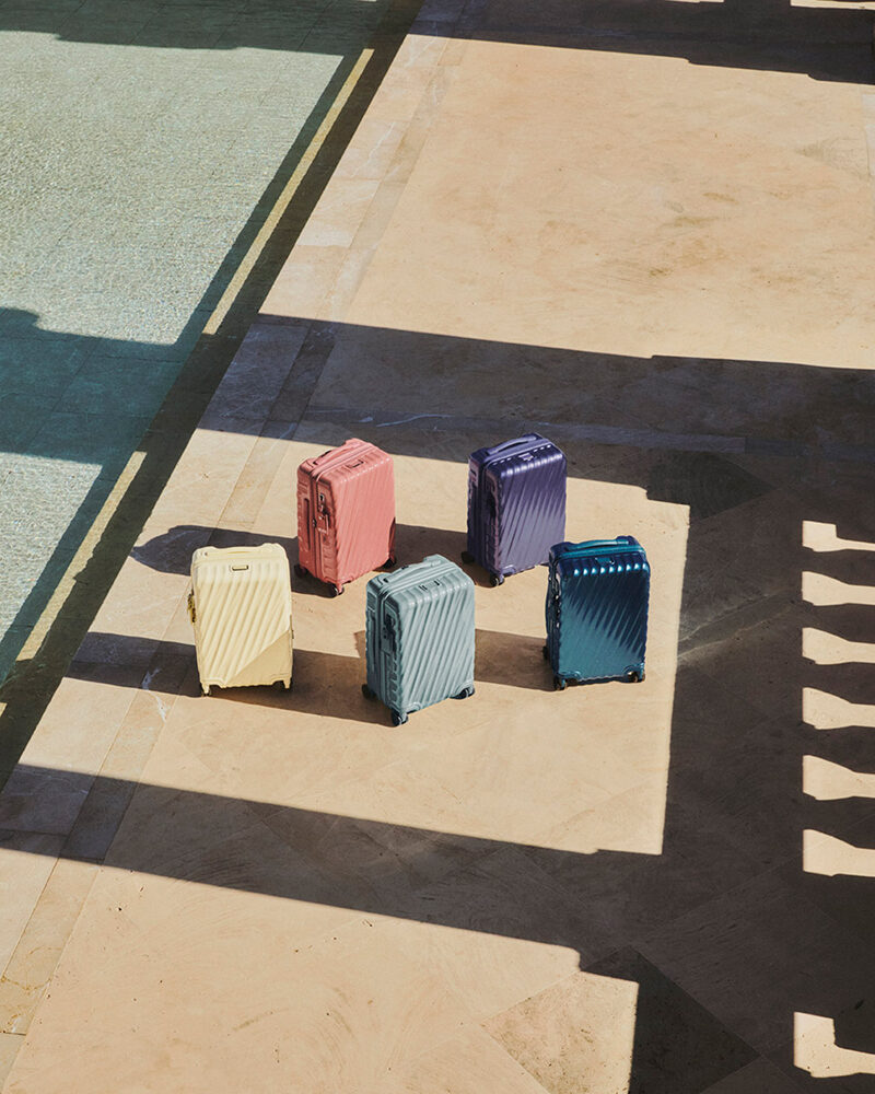 Mehrere kleine Koffer in Beige-, Blau-, Rosa- und Lilatönen stehen auf hellem Boden im Sonnenlicht.