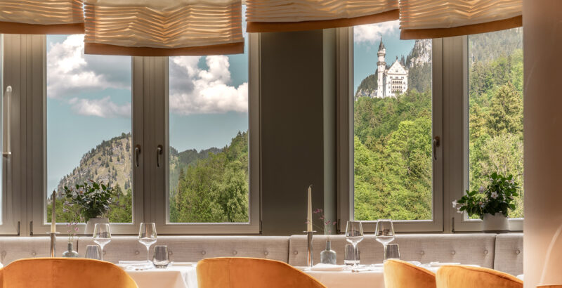 Heller Innenraum mit großen, rechteckigen Fenstern; davor eine lange Holzfläche oder Sitzbank, durch die Fenster Blick auf grüne Hügel und ein Schloss in der Ferne.