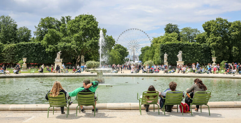 Grüne Metallstühle stehen rund um ein Wasserbecken in einer Parkanlage; im Hintergrund sind Springbrunnen und Bäume zu sehen.