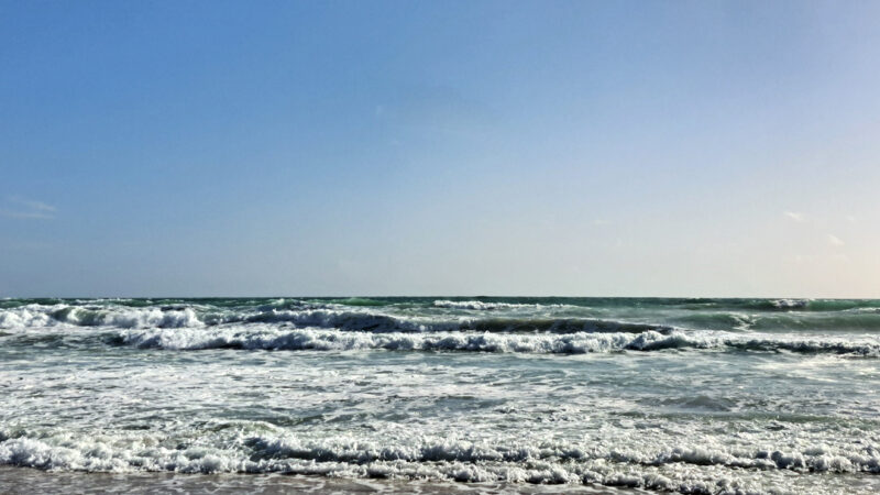 Weite Aufnahme eines Meeresstrandes mit flachen Wellen unter klarem Himmel; Horizont verläuft mittig durch das Bild.