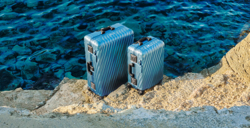 Zwei blaue Hartschalenkoffer stehen auf hellem Felsboden direkt vor dunkelblauem Wasser.