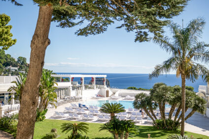 Blick durch Bäume und über Liegestühle am Pool auf das Meer und die Küstenlandschaft.