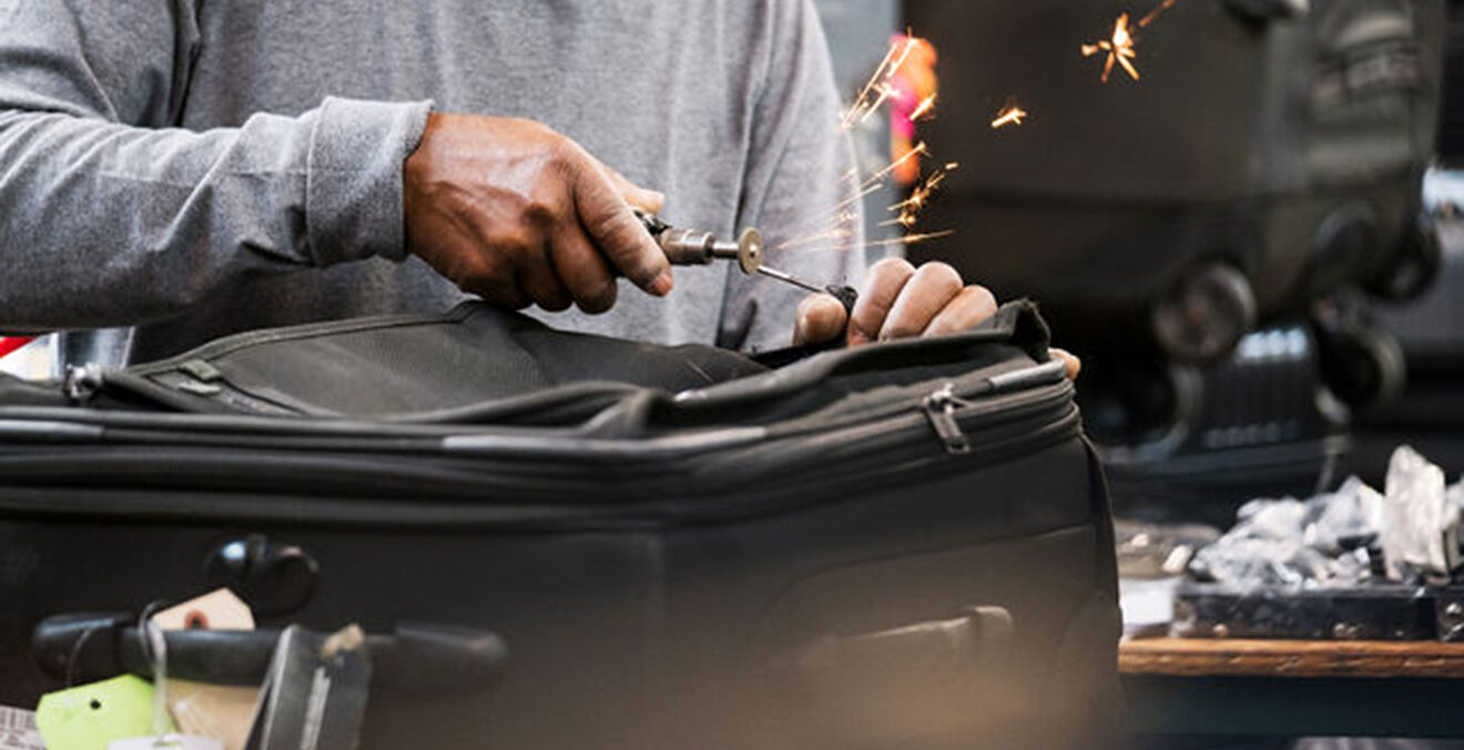 Nahaufnahme von Händen, die einen alten, schwarzen Koffer reparieren.