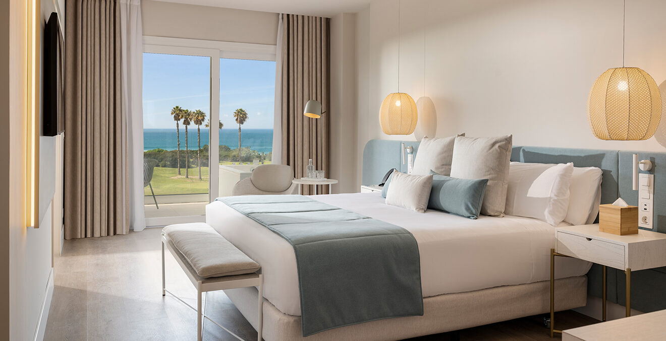 Zimmer mit großem Bett, weiß-blauen Kissen und Decken, großem Fenster mit Blick auf das Meer und Palmen im Hotel Amàre.
