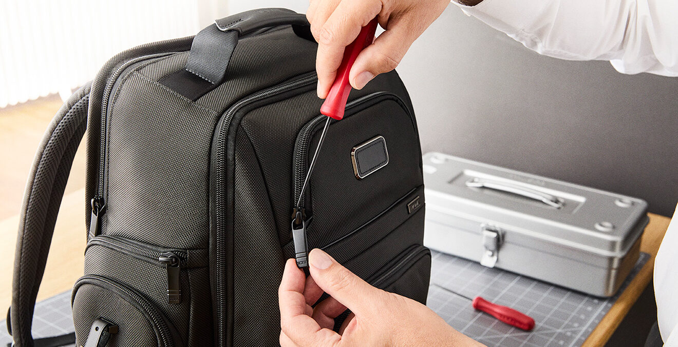 Nahaufnahme von Händen, die mit einem roten Werkzeug einen schwarzen Rucksack reparieren.
