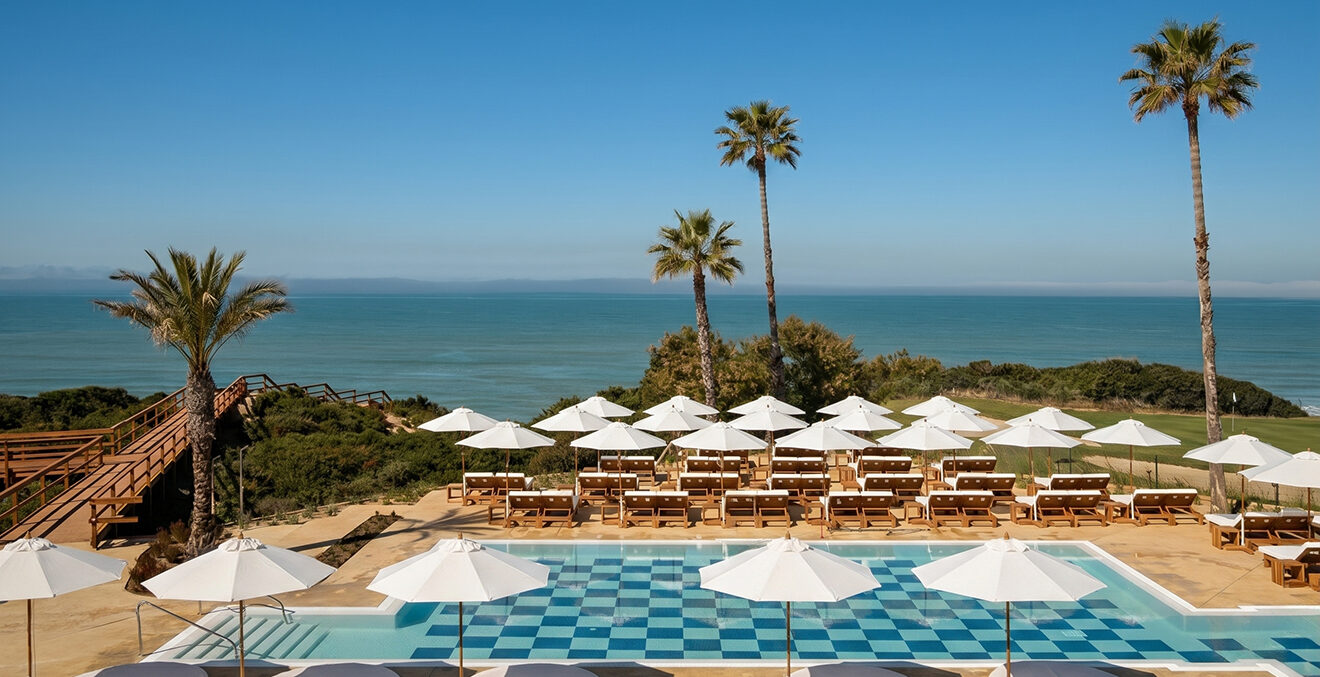Außenpool mit weißen Sonnenschirmen und Palmen sowie Blick auf das Meer.