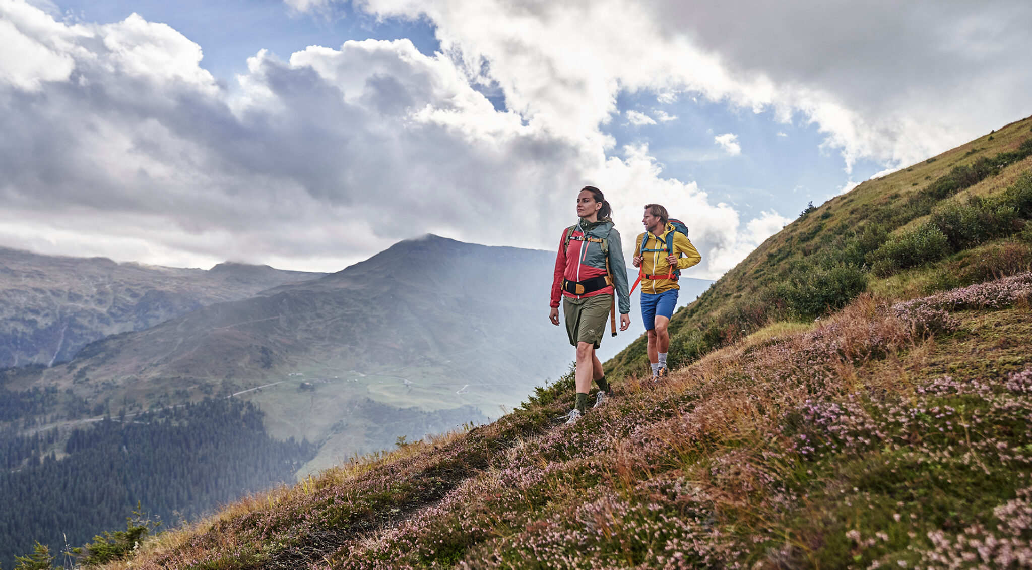 Zwei Wanderer mit Trekkingrucksäcken gehen über einen blühenden Berghang. Im Hintergrund erstreckt sich eine weite Gebirgslandschaft mit tief eingeschnittenen Tälern unter einem bewölkten Himmel.