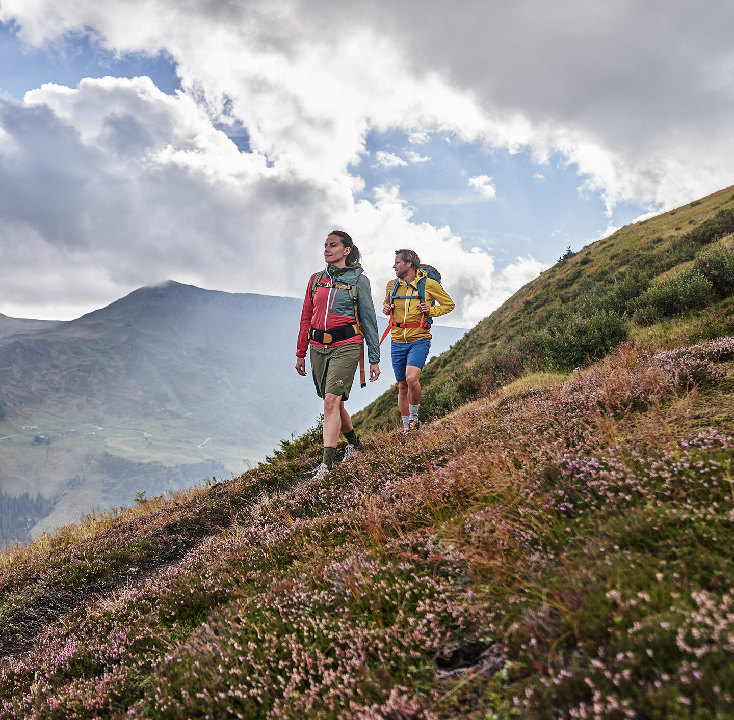 Zwei Wanderer mit Trekkingrucksäcken gehen über einen blühenden Berghang. Im Hintergrund erstreckt sich eine weite Gebirgslandschaft mit tief eingeschnittenen Tälern unter einem bewölkten Himmel.