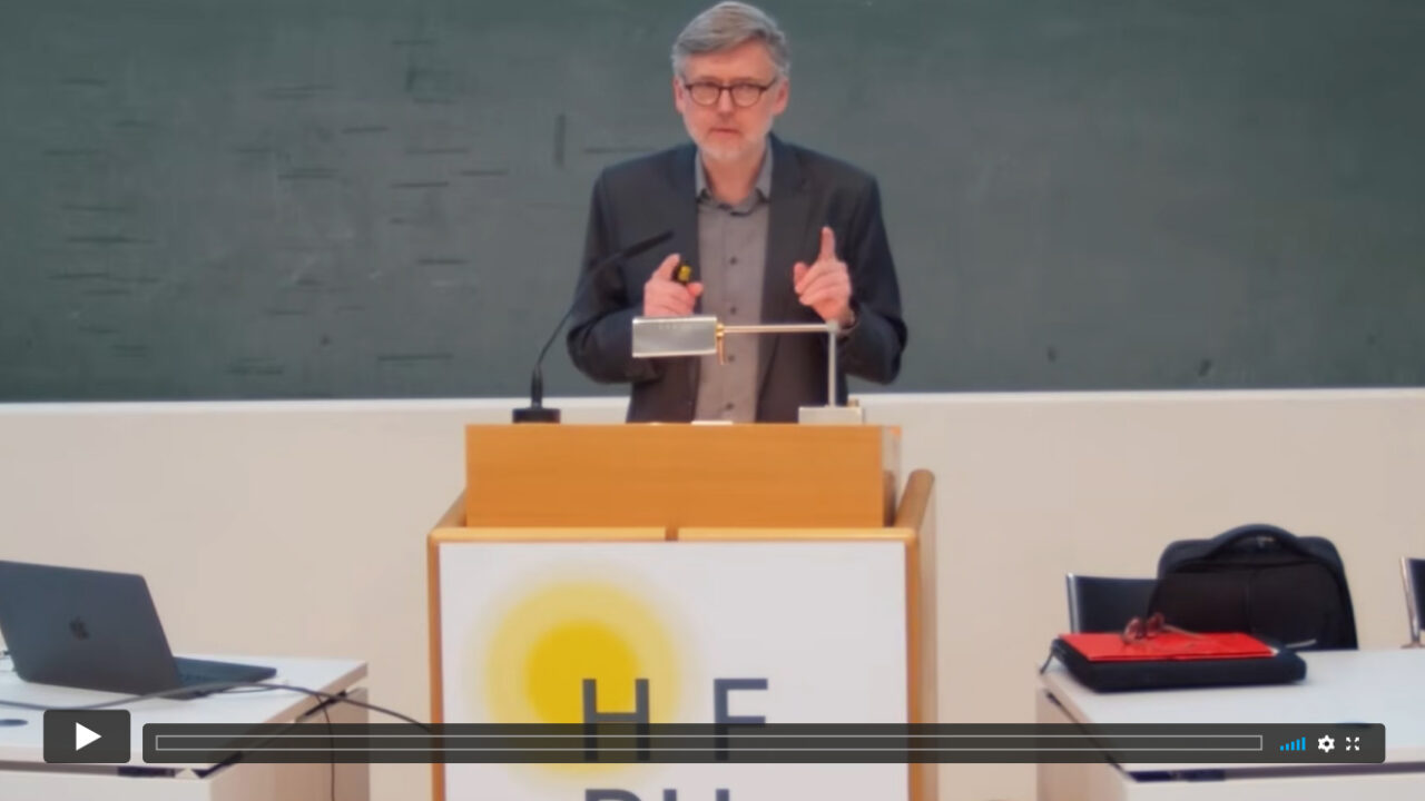 Dominik Finkelde SJ. steht hinter einem Rednerpult mit Mikrofon und gestikuliert während eines Vortrags. Im Hintergrund befindet sich eine grüne Tafel; auf der Vorderseite des Pults ist das Logo der Hochschule für Philosophie München zu sehen.