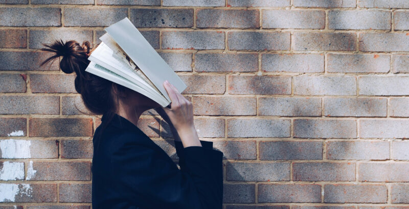 Eine Frau in dunkler Kleidung steht vor einer hellen Backsteinmauer. Ein aufgeschlagenes Buch bedeckt Kopf und Gesicht; die dunklen Haare trägt sie zu einem Dutt.