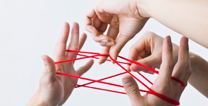 Mehrere Hände verschiedener Personen sind mit einem roten Faden verbunden. Die Finger halten und spannen die Fäden, sodass ein dichtes Netz zwischen den Händen entsteht.