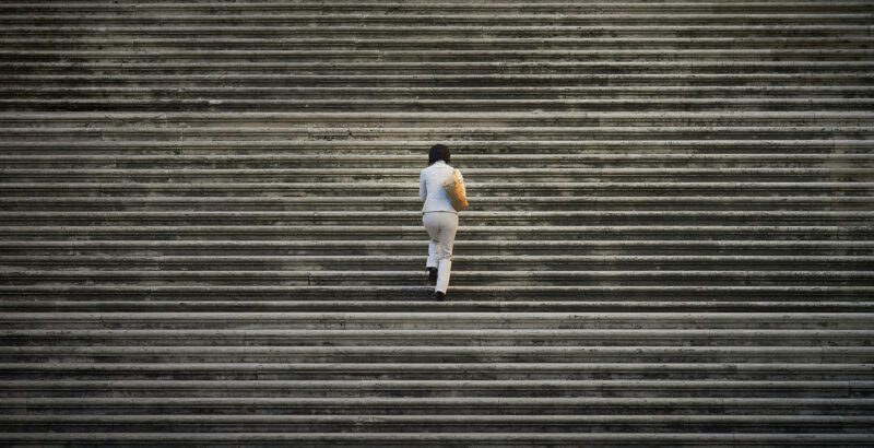 Luftaufnahme einer einzelnen Person in heller Kleidung, die mittig über breite, parallel verlaufende Betonstufen geht. Die regelmäßigen Linien der Treppenstufen füllen das gesamte Bild.