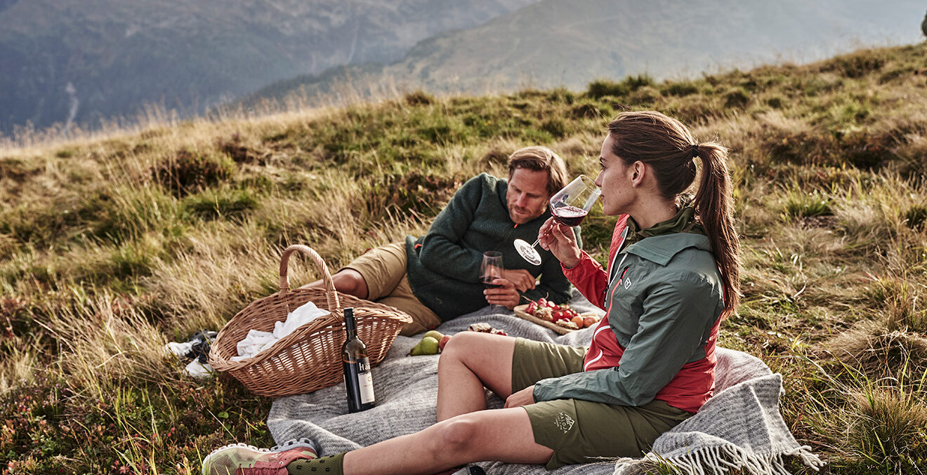 Ein Paar in Wanderkleidung macht ein Picknick auf einer Wiese vor Bergkulisse.