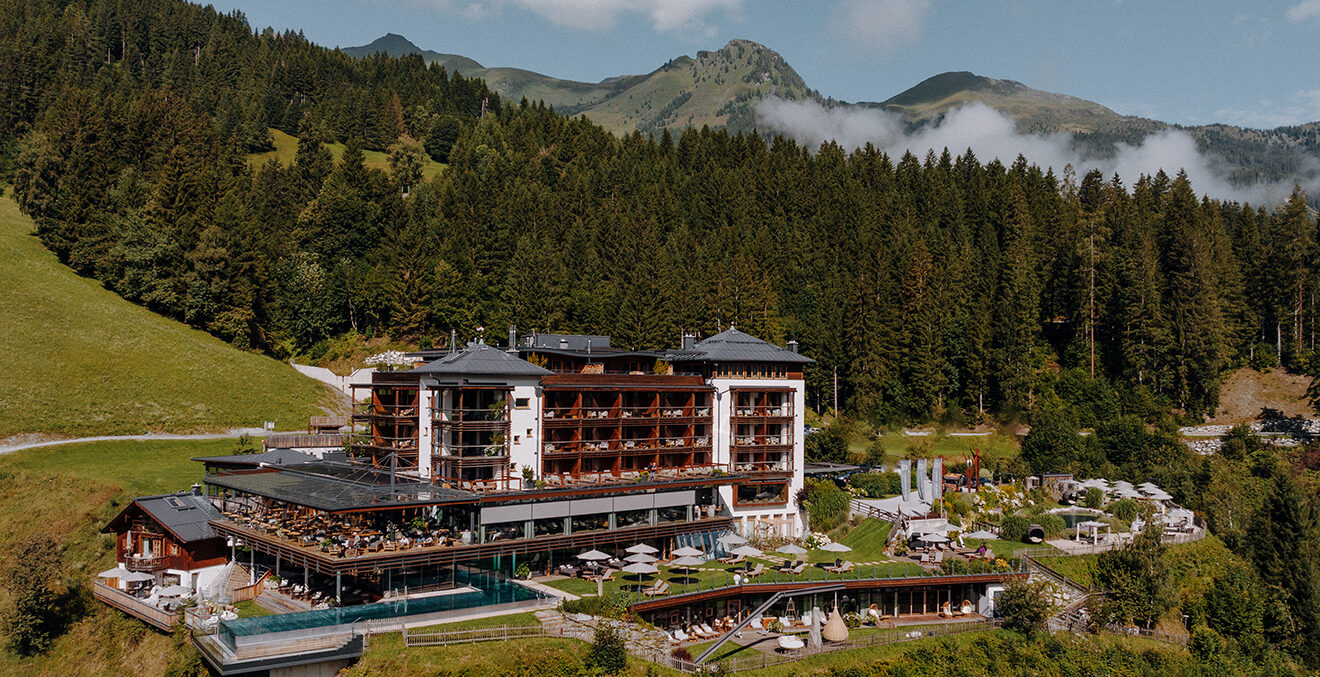 Mehrstöckiges Hotel mit Holzfassade und großen Glasflächen liegt eingebettet zwischen Wiesen und dichtem Nadelwald. Im Vordergrund befinden sich Außenpool, Sonnenterrassen und Liegebereiche; dahinter ragen bewaldete Berge auf.