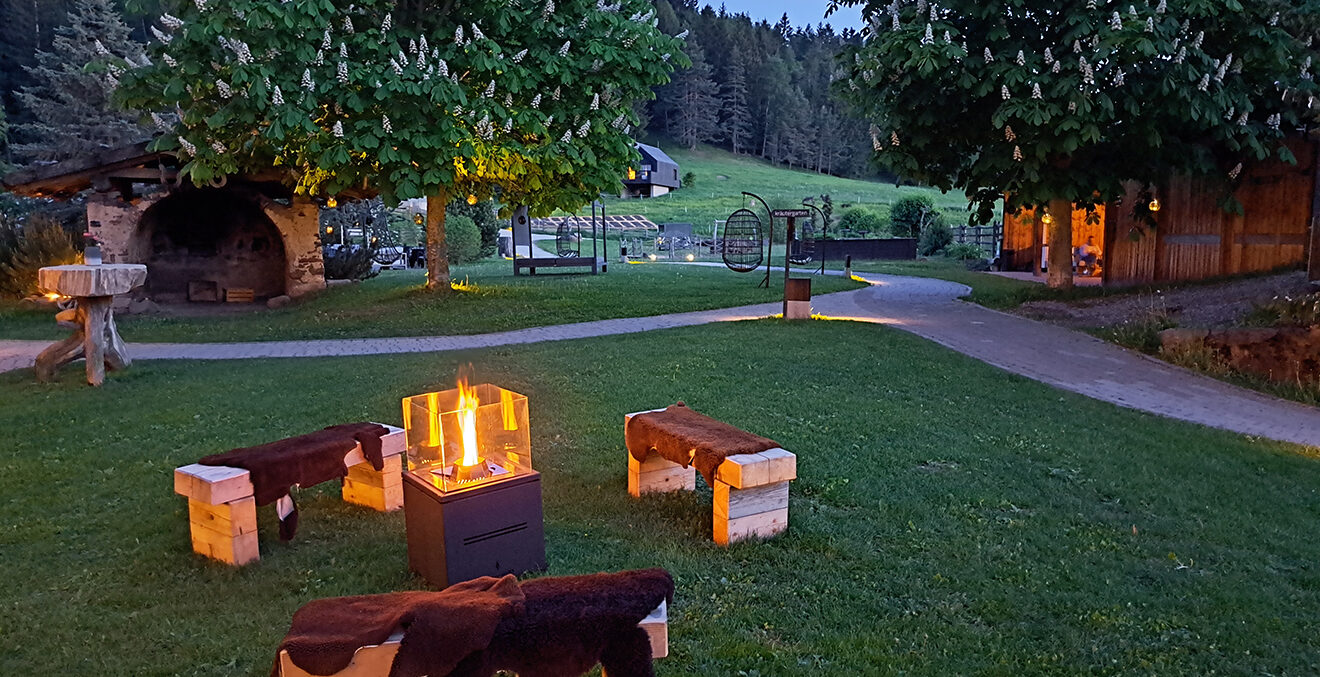 Abendstimmung mit schlichten Holzbänken auf einer grünen Wiese um eine Feuerstelle.