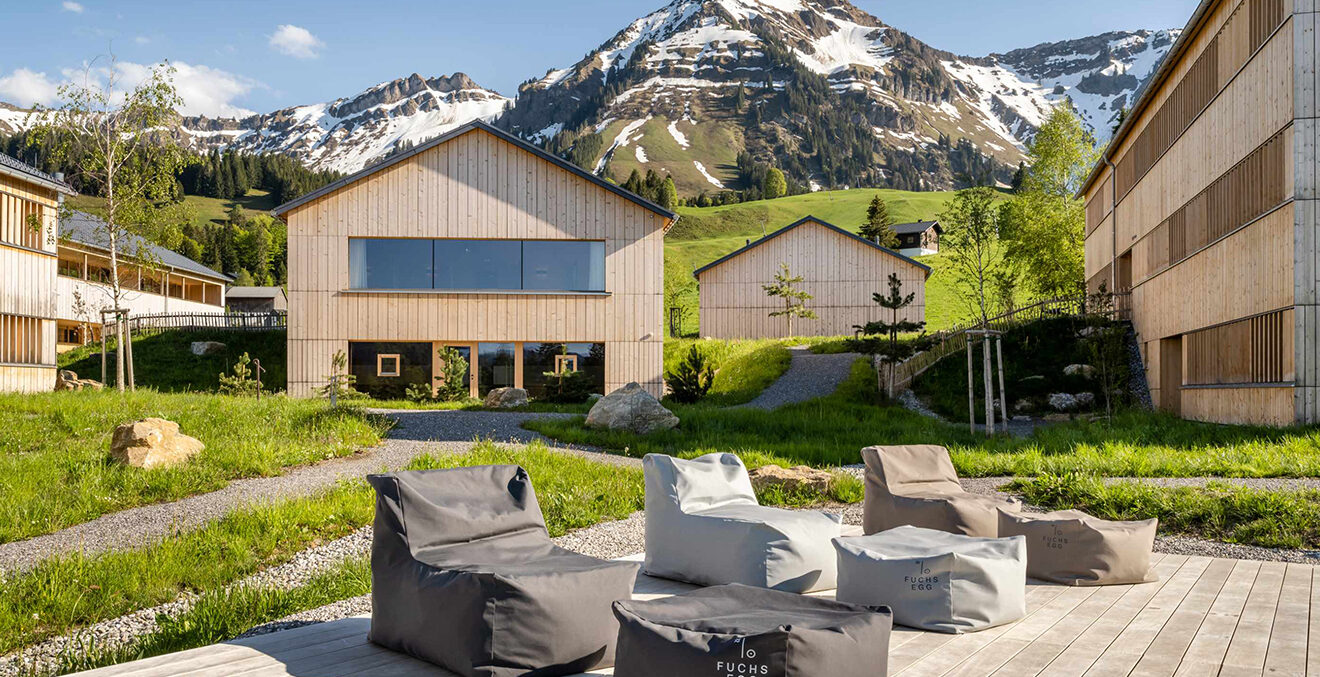 Moderner Innenhof mit gepolsterten Loungemöbeln auf einer Terrasse. Umgeben von Gebäuden mit Holzfassaden öffnet sich der Blick auf grüne Wiesen und eine Bergkulisse im Hintergrund.