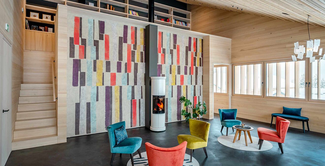 Mit hellem Holz und bunten Wandpaneelen ausgestatteter Raum. Bunte Sessel stehen um einen kleinen Ofen.