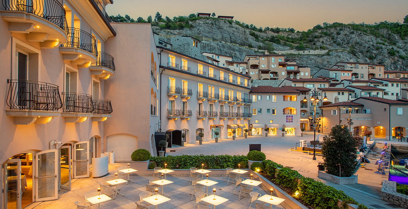 Moderne Hotel- und Apartmentanlage mit mehreren Stockwerken, Balkonen und großen Fensterflächen. Die Gebäude sind in der Abendstimmung warm beleuchtet; im Vordergrund liegt ein heller Platz mit Pool- oder Wasserfläche, im Hintergrund steigt ein dicht bebauter Hang an.