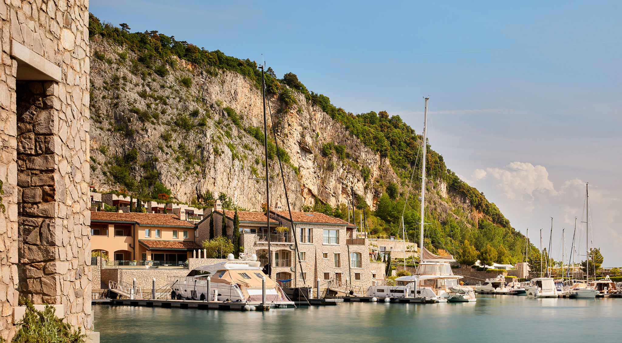 Panorama einer felsigen Adriaküste mit heller Kalksteinwand, mediterraner Vegetation und einem kleinen Yachthafen. Rechts liegen mehrere Segelboote mit hohen Masten im ruhigen Wasser, links ist eine Natursteinmauer zu sehen.