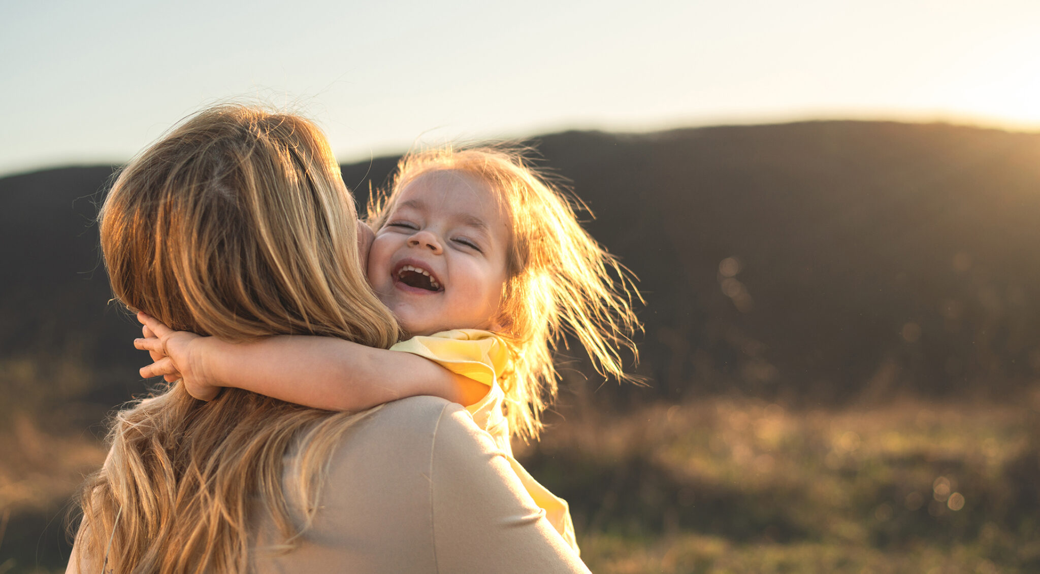 Nahaufnahme einer Mutter und ihrer kleinen Tochter im warmen Gegenlicht. Das Kind hat die Augen geschlossen und lacht, während es von der Mutter eng umarmt wird; beide Gesichter berühren sich, im Hintergrund liegt eine unscharfe Landschaft.