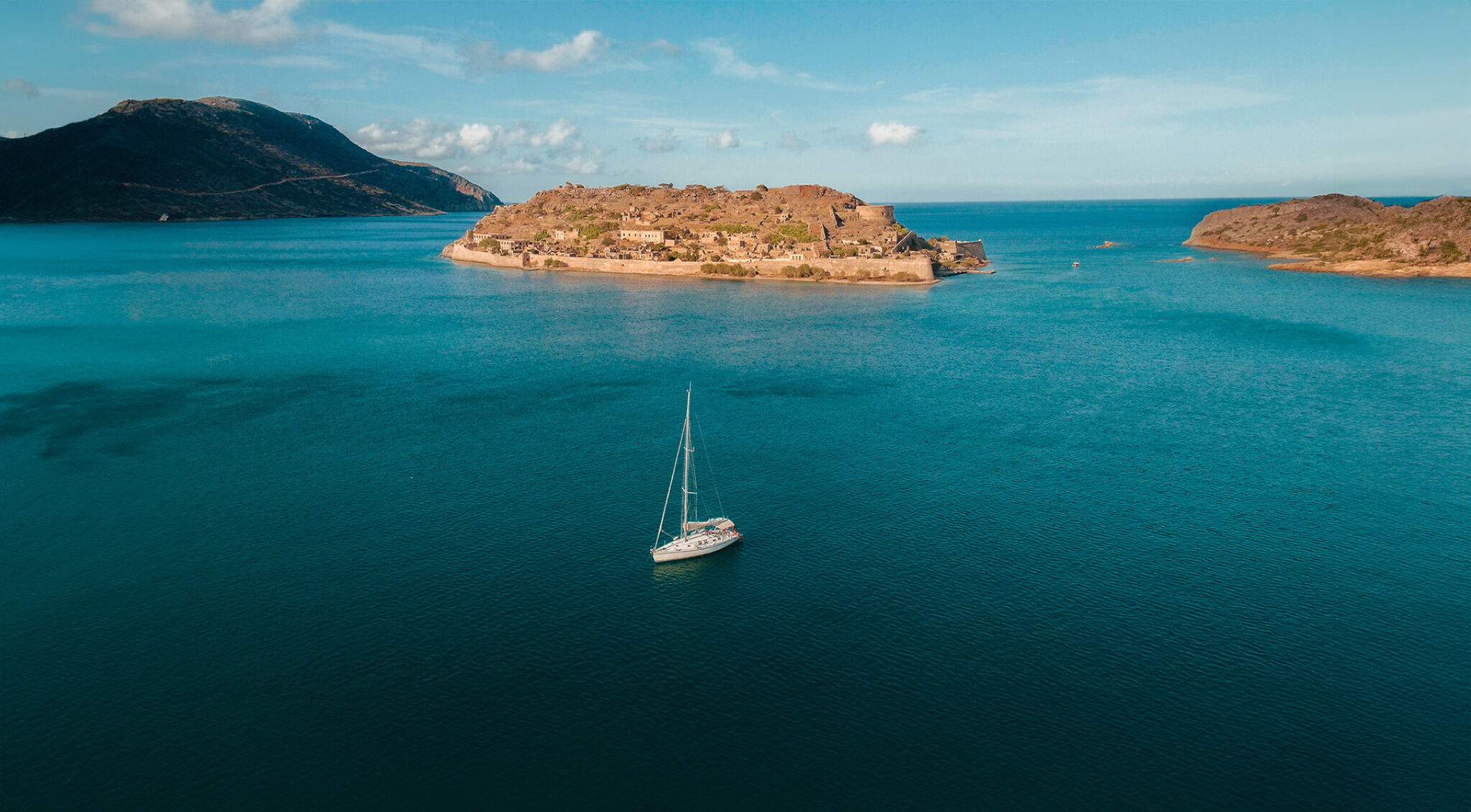 Luftaufnahme einer weiten Bucht auf Kreta mit tiefblauem Meer. In der Bildmitte liegt eine felsige, dicht bebaute Insel mit historischen Gebäuden und Mauern; davor segelt ein weißes Segelboot. Links und rechts rahmen bergige, karge Küstenabschnitte die Bucht.