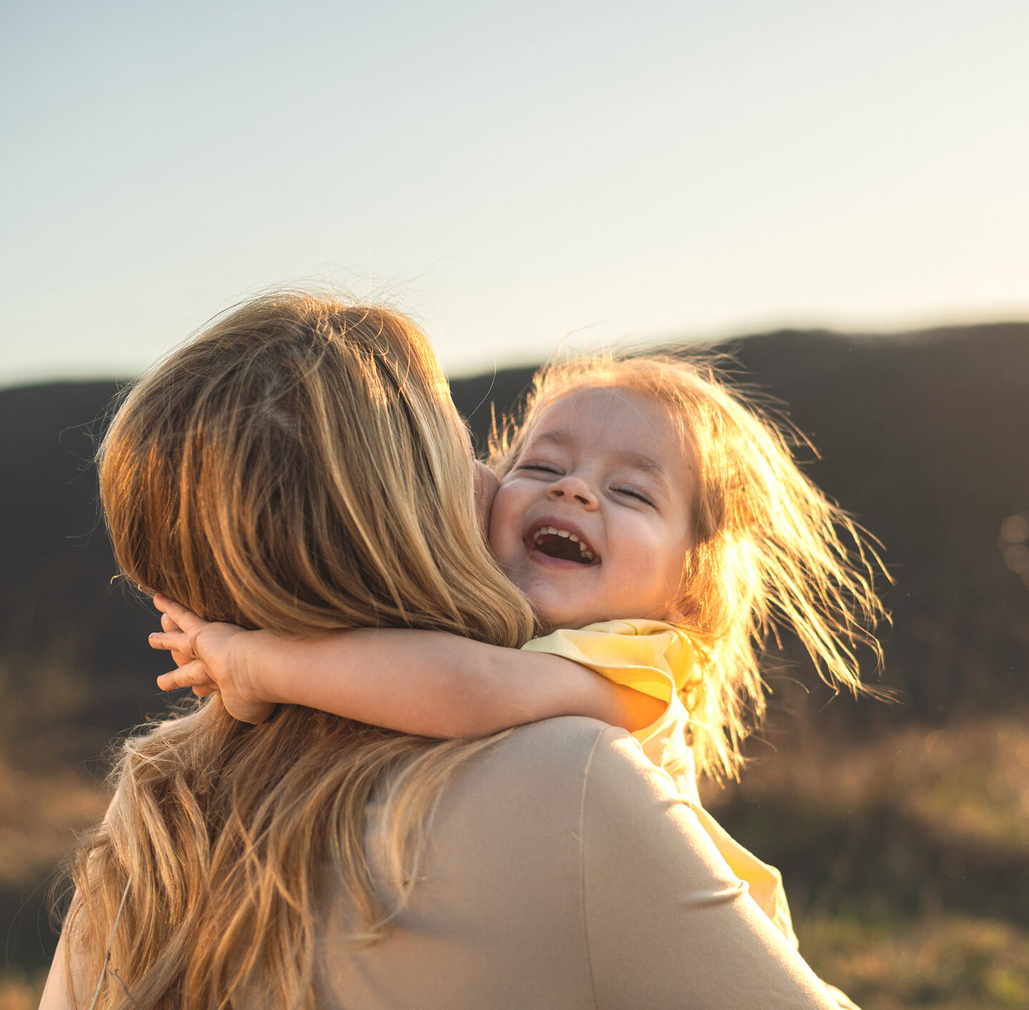 Nahaufnahme einer Mutter und ihrer kleinen Tochter im warmen Gegenlicht. Das Kind hat die Augen geschlossen und lacht, während es von der Mutter eng umarmt wird; beide Gesichter berühren sich, im Hintergrund liegt eine unscharfe Landschaft.