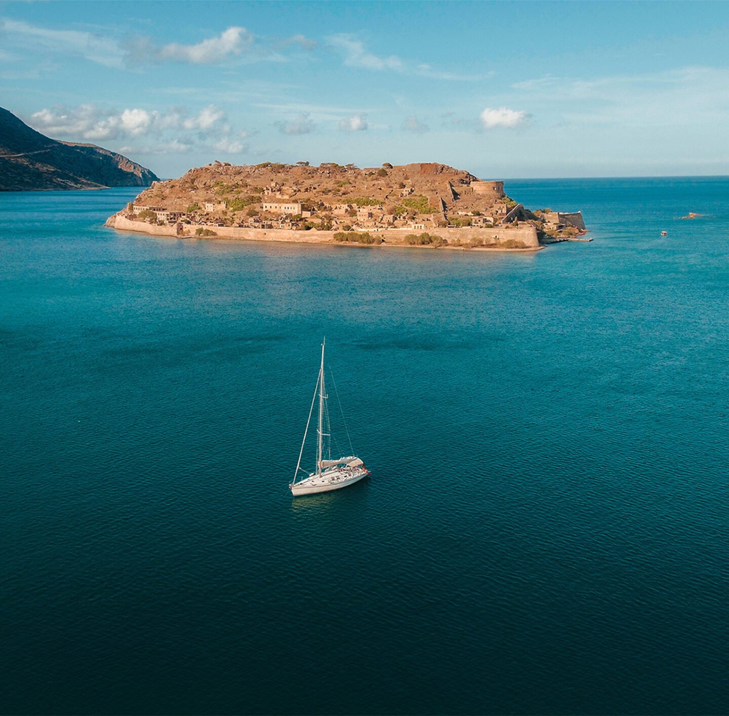 Luftaufnahme einer weiten Bucht auf Kreta mit tiefblauem Meer. In der Bildmitte liegt eine felsige, dicht bebaute Insel mit historischen Gebäuden und Mauern; davor segelt ein weißes Segelboot. Links und rechts rahmen bergige, karge Küstenabschnitte die Bucht.