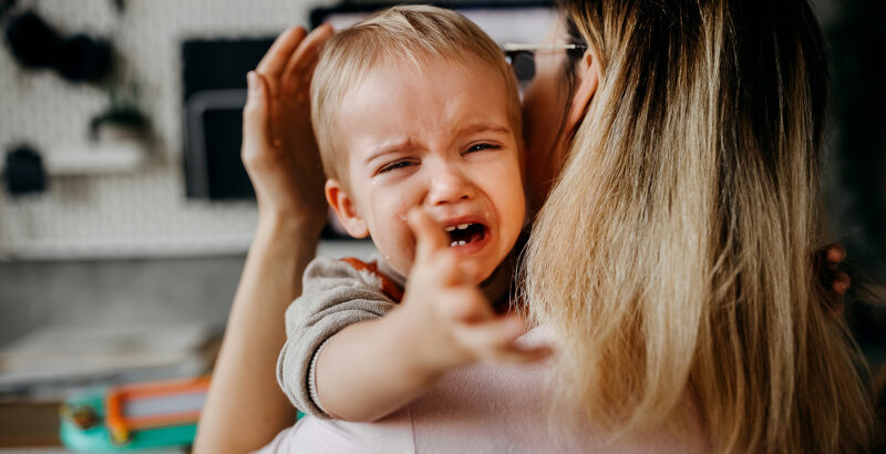 Nahaufnahme eines kleinen Kindes auf dem Arm einer Frau. Das Kind weint oder ruft mit geöffnetem Mund und ausgestreckter Hand; rechts sind die langen blonden Haare und die Schulter der Frau zu sehen.
