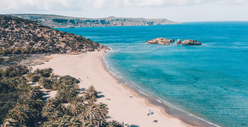 Luftaufnahme eines langen, hellen Sandstrandes an türkisblauem Meer. Links grenzt ein dichter Palmenhain an den Strand, auf dem vereinzelt Menschen und Strandliegen zu erkennen sind.
