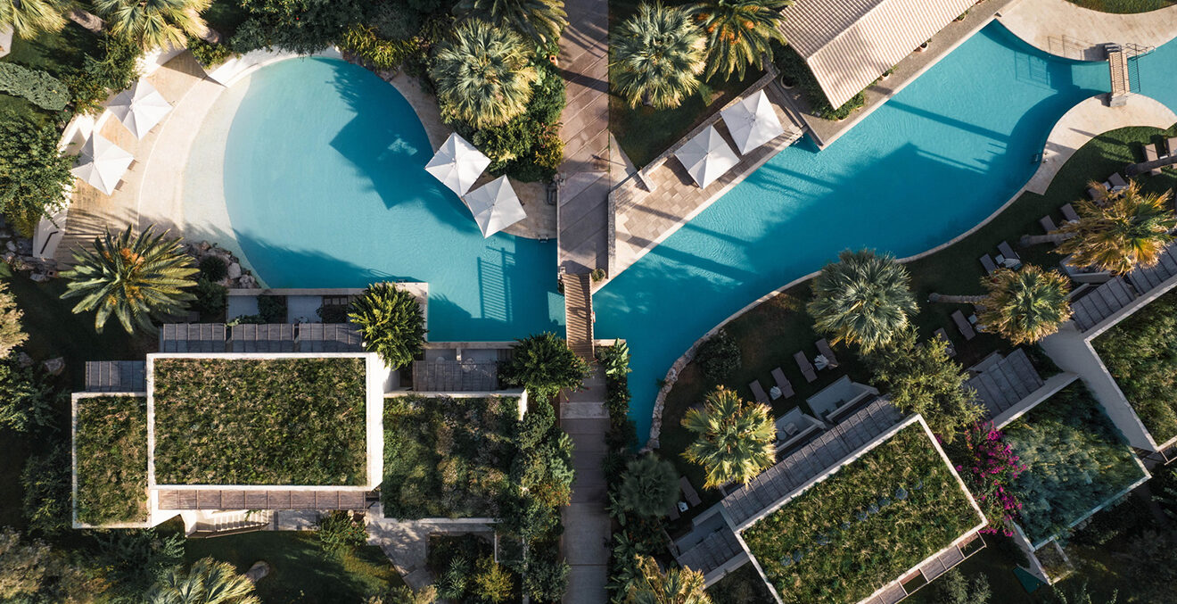 Senkrechte Luftaufnahme der Poolanlage des Phāea Blue mit mit zwei geschwungenen türkisfarbenen Pools. Weiße Sonnenschirme, Liegen, Palmen und Wege liegen zwischen Gebäuden mit begrünten Flachdächern.
