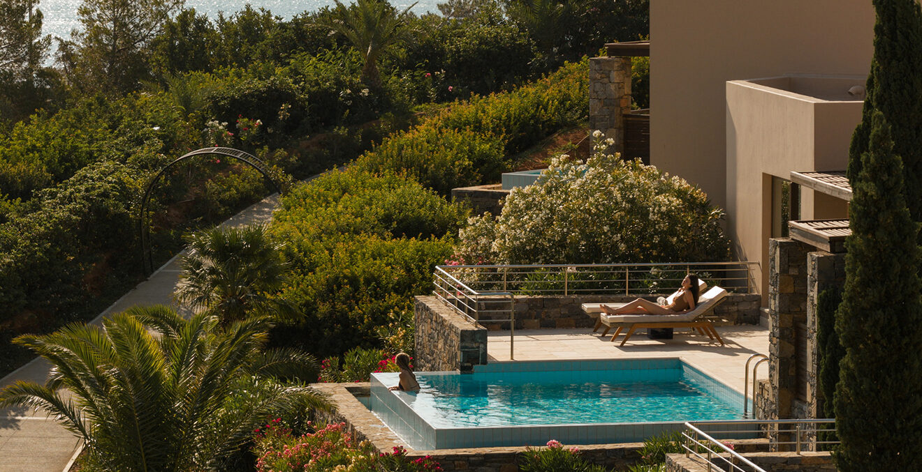 Kleiner rechteckiger Privatpool vor einer modernen, beigefarbenen Hotelvilla. Eine Person liegt auf einer Sonnenliege neben dem Pool; die Terrasse ist von dichtem mediterranem Grün, Palmen und blühenden Sträuchern umgeben.