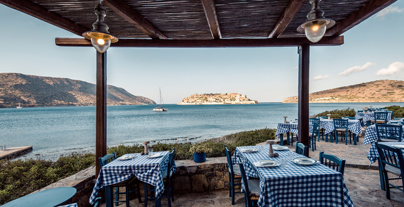 Restaurantterrasse mit Tischen gedeckt mit blau-weißen Tischdecken und Blick in eine Bucht.