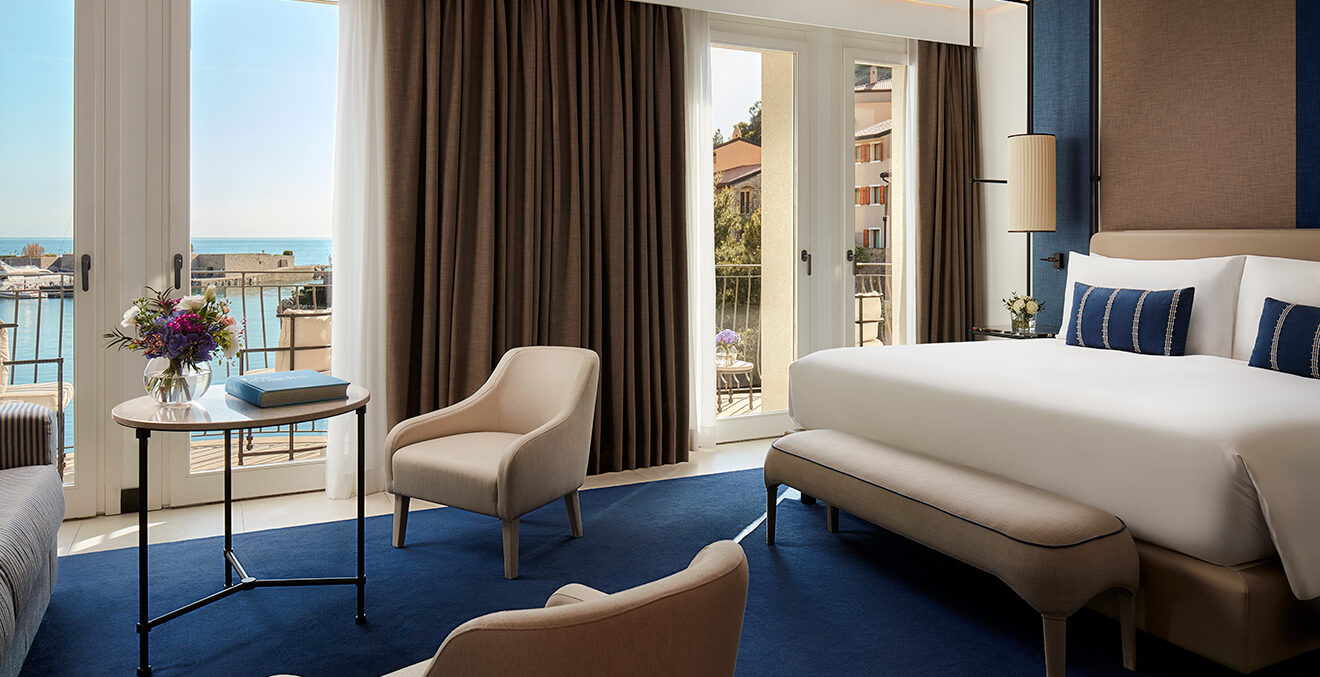 Hotelzimmer Portopiccolo mit braunem Vorhang, dunkelblauem Teppich und hellem Mobiliar.