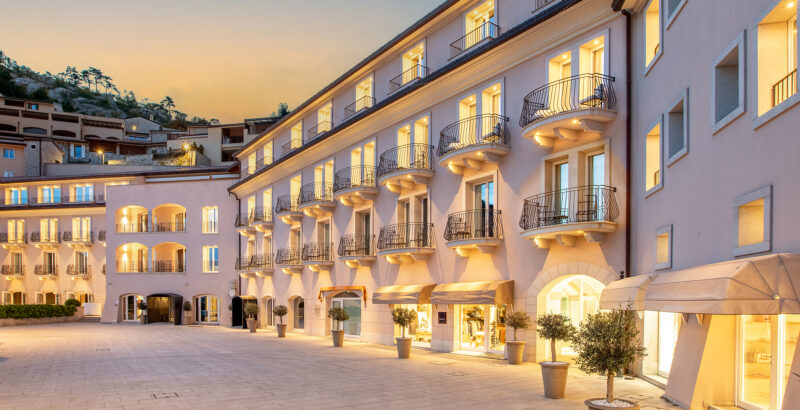 Breite Ansicht einer modernen Wohn- oder Hotelanlage mit mehreren Etagen, vielen Balkonen und großen Glasflächen. Die Fassade ist in hellen Grau- und Beigetönen gehalten und am Abend warm beleuchtet.