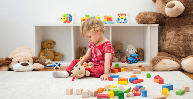 Blondes Kleinkind in rotem Oberteil sitzt auf einem hellen Teppich und spielt mit bunten Holzbausteinen. Um das Kind liegen zahlreiche farbige Klötze, Tierfiguren und Spielsachen; im Hintergrund sitzen große Stofftiere vor einem Regal.
