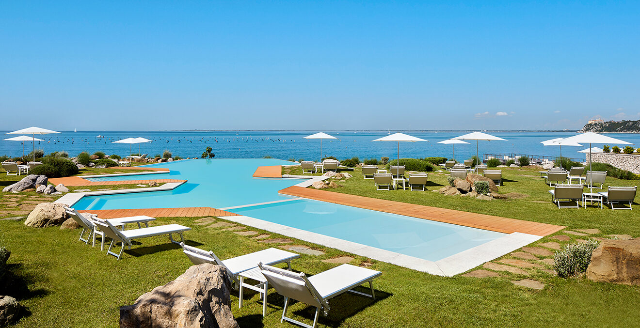 Poolanlage mit Blick auf das Meer und weißen Liegestühlen auf grüner Wiese.