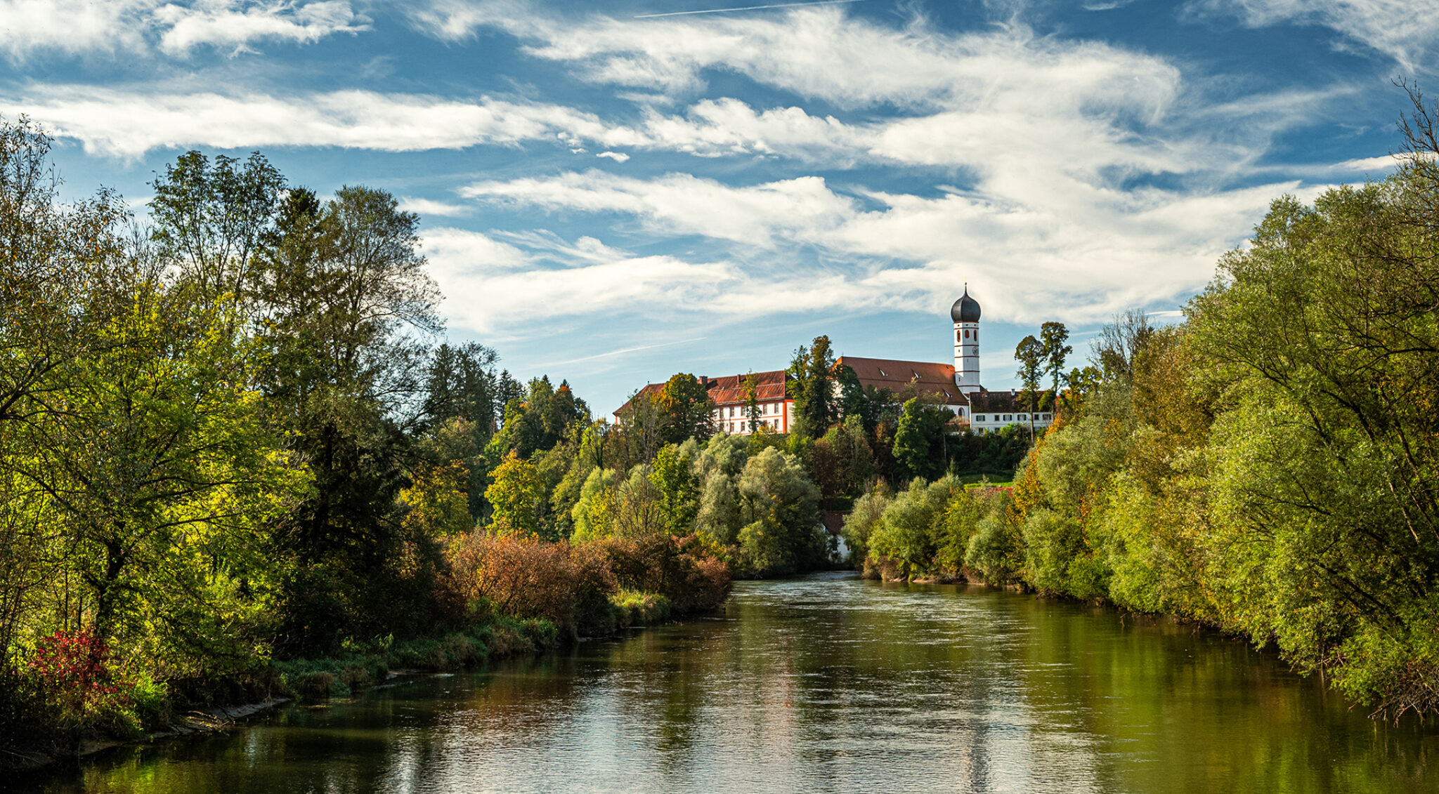Panoramaaufnahme der Klosteranlage Beuerberg auf einer Anhöhe. Im Vordergrund erstrecken sich Wiesen und Bäume im frischen Grün, im Hintergrund stehen mehrere historische Gebäude mit roten Dächern unter einem blau-weiß bewölkten Himmel.