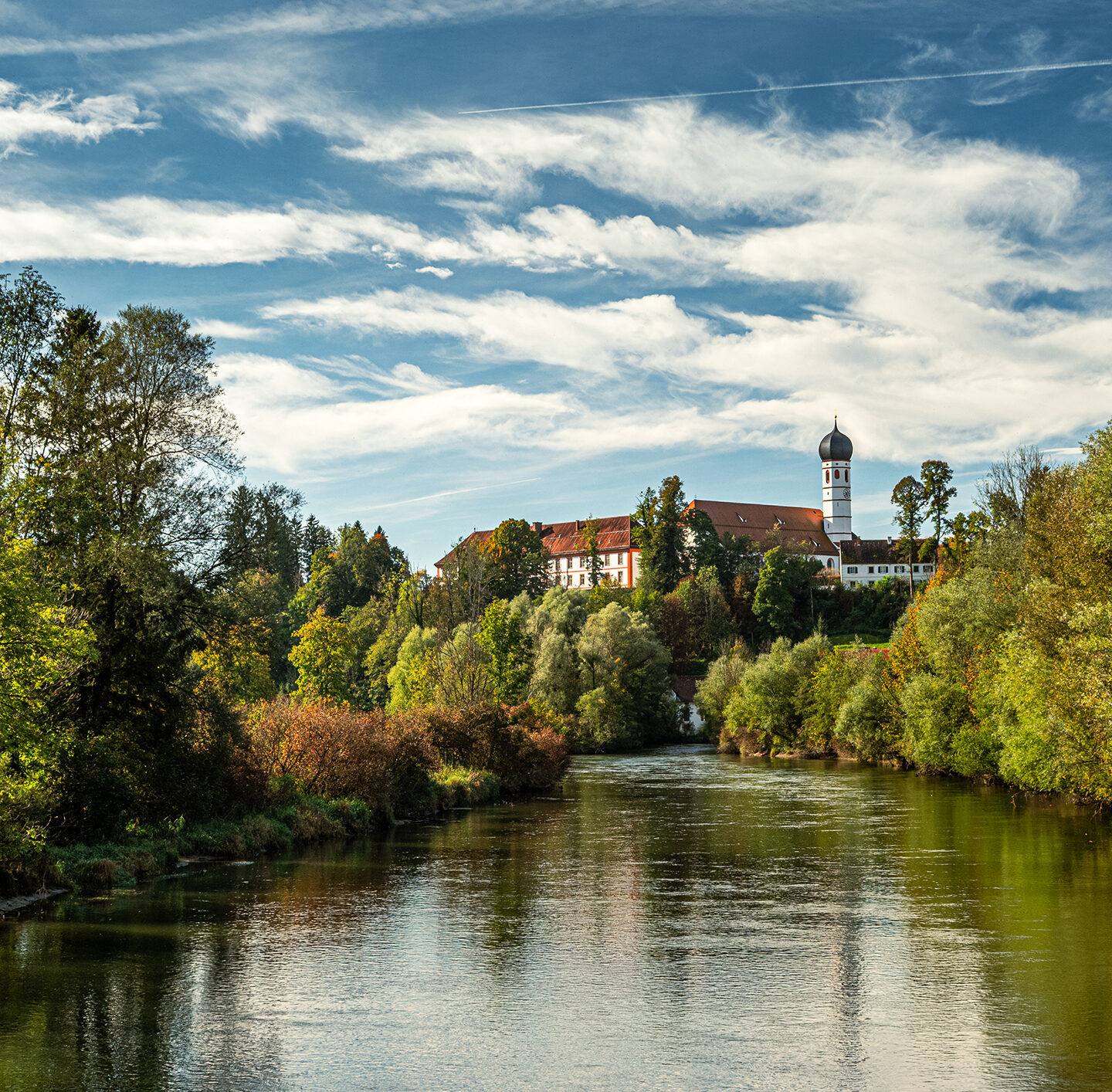 Panoramaaufnahme der Klosteranlage Beuerberg auf einer Anhöhe. Im Vordergrund erstrecken sich Wiesen und Bäume im frischen Grün, im Hintergrund stehen mehrere historische Gebäude mit roten Dächern unter einem blau-weiß bewölkten Himmel.