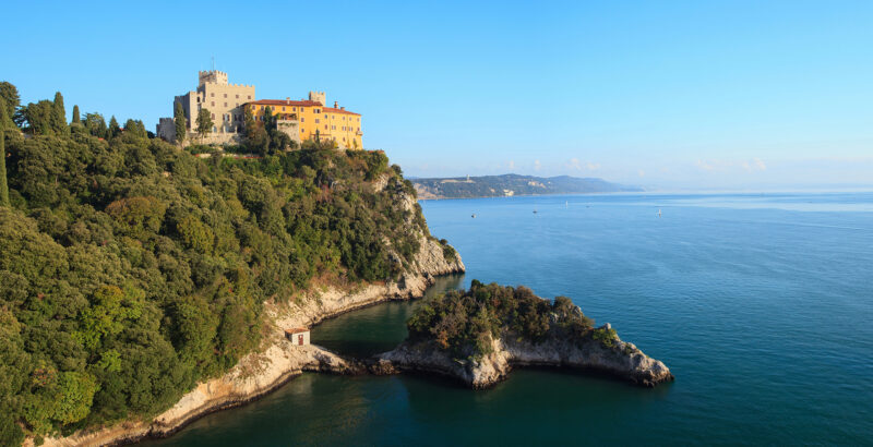 Blick auf Schloss Duino; Auf dem erhöhten Plateau steht eine längliche helle Festungs- oder Gebäudestruktur; rechts öffnet sich das ruhige blaue Meer bis zum Horizont.